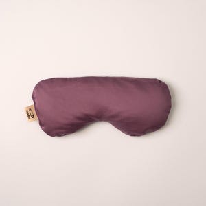 May include: A plum-colored sleep mask with a smooth, satin-like finish and a contoured shape. A small, light-colored fabric tag is attached to the left side, featuring a black safety pin icon and the text MARV printed in a simple, bold font.