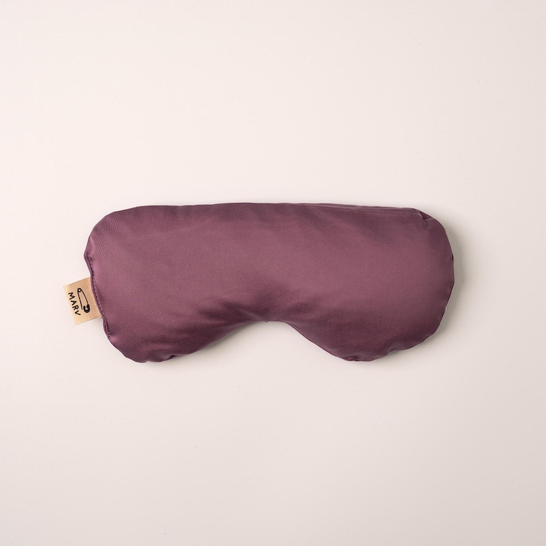 May include: A plum-colored sleep mask with a smooth, satin-like finish and a contoured shape. A small, light-colored fabric tag is attached to the left side, featuring a black safety pin icon and the text MARV printed in a simple, bold font.