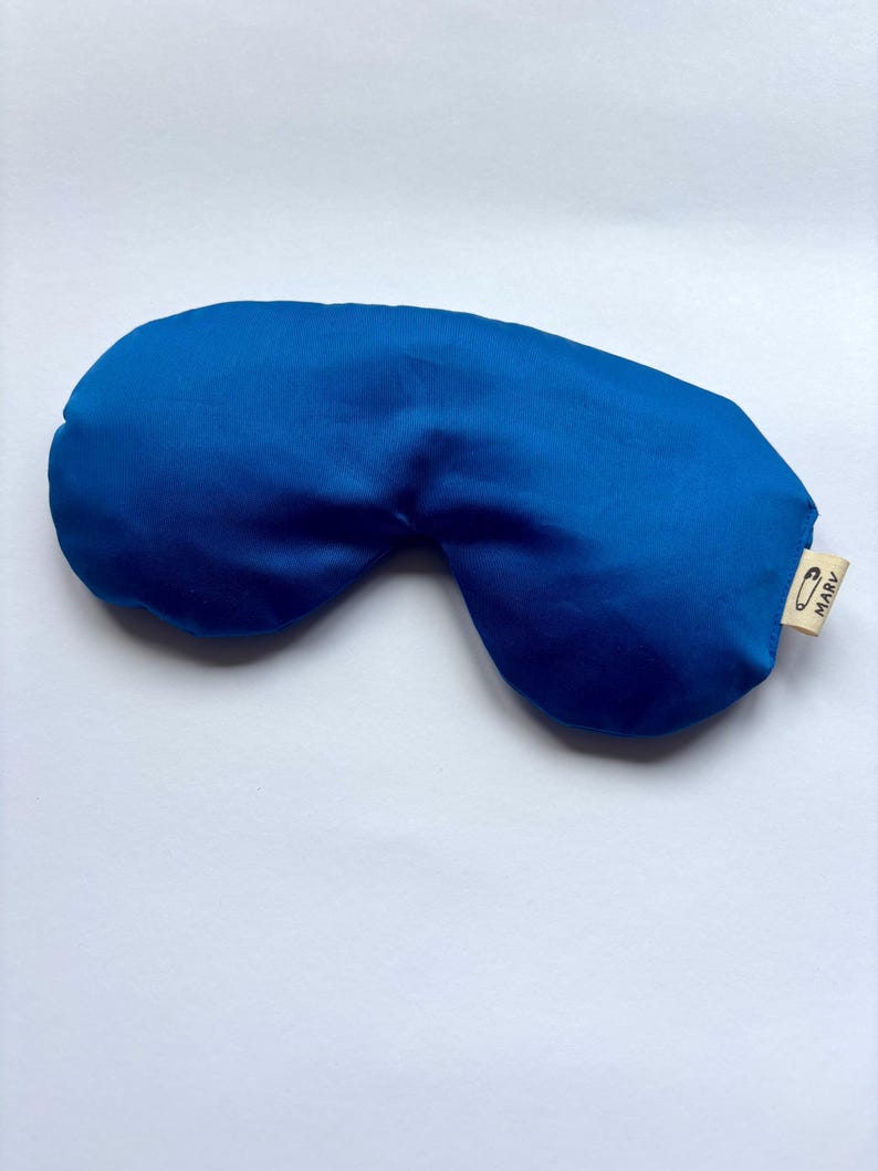 May include: A solid royal blue sleep mask with a smooth, satin-like finish. The mask features a small, light beige fabric tag on the right side, which displays a black safety pin icon and the text MARV printed in a clear, sans-serif font.