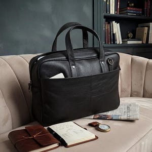 May include: A black leather briefcase with two top handles, a front slip pocket, and silver-toned hardware. The bag features a textured grain finish, a zippered main compartment, and side D-rings for an optional shoulder strap attachment.