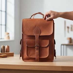 May include: A brown leather backpack with a flap closure, two front pockets, and a top handle. The bag has a buckle closure and a shoulder strap. The bag is sitting on a wooden surface.