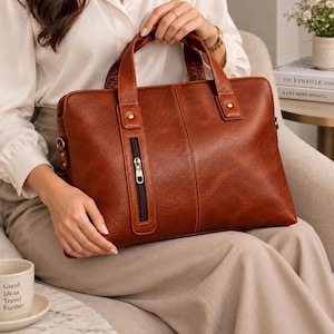 May include: A structured cognac brown leather briefcase featuring two top handles, a vertical zippered front pocket with a metallic pull, and reinforced stitching details. The bag includes side hardware for an optional shoulder strap attachment.