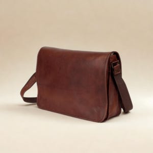 May include: A dark brown leather messenger bag featuring a rectangular flap closure, a smooth finish, and a wide adjustable fabric shoulder strap attached to the sides with metal hardware. The bag has a structured design with visible stitching.