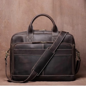 May include: A dark brown leather briefcase featuring two top handles, a detachable shoulder strap with a padded section, and a front zippered pocket. The bag is accented with visible white contrast stitching and antique brass-toned metal hardware.