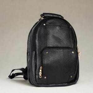 May include: Black leather backpack with a rounded top, a front pocket, and a top handle. The backpack has gold-colored zippers and rivets. The shoulder straps are visible on the left side of the image.