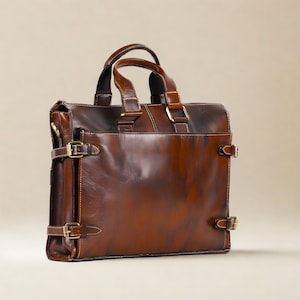 May include: A structured brown leather briefcase featuring a polished finish with darker shaded areas. The bag includes two top handles, a front pocket, and side straps secured with brass-toned metal buckles for a classic, functional design.