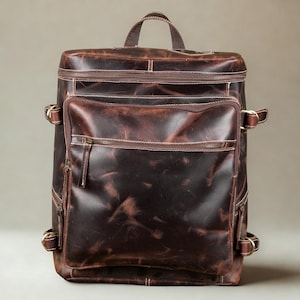 Puede incluir: Mochila de cuero marrón con múltiples compartimentos con cremallera y correas ajustables. La mochila tiene un asa superior y un acabado envejecido, con costuras visibles y herrajes metálicos. La bolsa es de forma rectangular.
