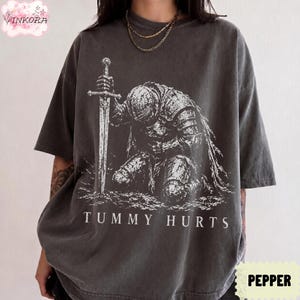 May include: A charcoal grey short-sleeve t-shirt featuring a white graphic print of a knight in armor kneeling and holding a sword. The text "TUMMY HURTS" is printed in white capital letters below the knight illustration on the front of the shirt.