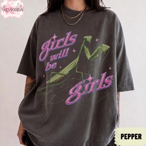 May include: A charcoal gray short-sleeve t-shirt featuring a green praying mantis graphic and the text "Girls will be girls" in a pink neon-style font. The design includes small pink star accents scattered around the insect and the lettering.