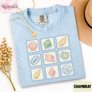 May include: Light blue t-shirt with a seashell stamp design. The shirt features nine different seashell illustrations in various colors. The shirt is made of a soft material and is labeled "CHAMBRAY".