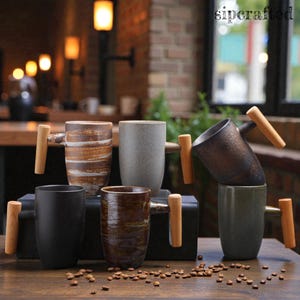 Könnte beinhalten: Eine Sammlung von Keramikbechern mit Holzgriffen. Die Becher sind in verschiedenen Farben erhältlich, darunter Schwarz, Braun, Grau und Grün, und weisen einzigartige strukturierte Muster auf. Die Becher werden auf einem Holztisch mit verstreuten Kaffeebohnen präsentiert.