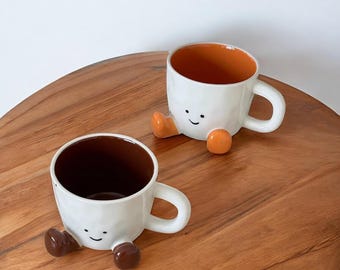 Peut inclure: Deux tasses en céramique blanche avec un motif de visage souriant et de petits pieds arrondis. L'une a un intérieur et des pieds orange, l'autre a un intérieur et des pieds marron. Les deux tasses ont une anse et sont sur une surface en bois.