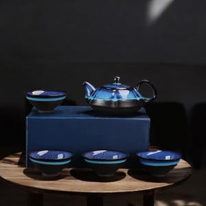 May include: A ceramic tea set with a blue and black glaze. The set includes a teapot and four matching teacups. The teapot has a black handle and lid. The teacups have a blue band around the rim. The set is displayed on a wooden table.