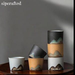 Può includere: Una collezione di tazze in ceramica bianche, nere e beige, ognuna con un design a catena montuosa marrone e verde. Le tazze sono impilate e disposte su una superficie scura. Il testo "sipcrafted" è visibile.