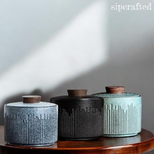 Pode incluir: Três potes de cerâmica com tampas de madeira. Os potes são cilíndricos, com linhas verticais texturizadas. As cores são azul-acinzentado, preto e verde-azulado. Os potes são exibidos em uma mesa de madeira escura. O texto "sipcrafted" está no canto superior direito.