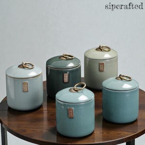 Op de afbeelding: Vijf keramische potten in blauw- en groentinten staan op een houten tafel. Elke pot heeft een deksel met een goudkleurige ringgreep en een klein rechthoekig etiket. De potten variëren in grootte en kleur, wat een aantrekkelijk geheel creëert.