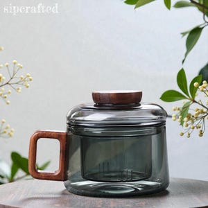May include: A glass tea mug with a wooden handle and lid. The mug features a built-in infuser and a contemporary design. The handle and lid are made of wood. The text "sipcrafted" is in the upper left corner of the image.