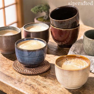 May include: A collection of ceramic coffee cups in various earth-tone colors, filled with coffee. The cups are placed on a wooden table, with one cup on a woven coaster. The image includes the text "sipcrafted".