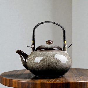 May include: A speckled ceramic teapot with a dark brown handle and spout. The teapot has a rounded body and a lid with a small knob. The handle is black and wrapped with a woven material. The teapot is sitting on a wooden table.