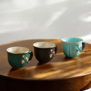 May include: Three ceramic teacups in teal, black, and green, each with a gold rim and handle. The cups feature gold floral accents and sit on a wooden surface. The teacups are designed for tea or coffee.