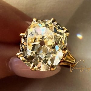 May include: A large, gold-toned ring featuring a prominent, faceted, light-yellow gemstone. The ring's band and setting are also gold. The gemstone is rectangular with rounded corners. The ring is held up by a person's hand.