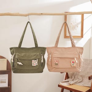 May include: Two corduroy tote bags, one olive green and one beige, hanging from a wooden branch. Each bag has a front pocket, zipper details, and decorative patches. The bags are suspended in front of a neutral-toned wall.