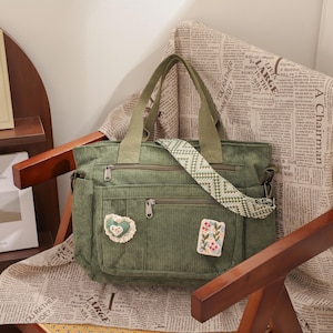 Pode incluir: Uma sacola tote de veludo cotelê verde com vários bolsos e uma alça estampada está em uma cadeira coberta com tecido com estampa de jornal. A bolsa tem um patch de coração e floral. Um livro intitulado "Industrial Drawing" é visível.