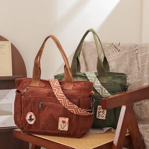 May include: Two corduroy tote bags in rust and olive green sit on a wooden chair. The rust-colored bag has a patterned strap and decorative patches. The olive green bag has a solid green strap. Both bags have top handles and exterior pockets.