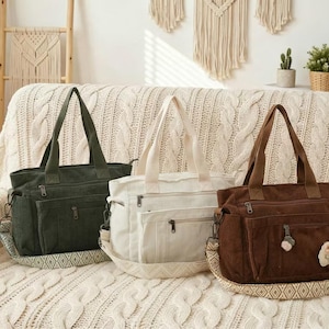 Può includere: Tre borse tote in tela verde, bianca e marrone. Ogni borsa ha una tracolla, più tasche e un cinturino decorativo fantasia. Le borse sono esposte su una superficie strutturata color crema.