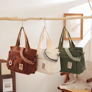 May include: Three canvas tote bags in brown, cream, and green hang from a wooden branch. The brown bag has a floral patch and patterned strap. The cream bag has a black and white embroidered trim. The green bag has a floral patch and patterned strap.