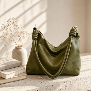 Puede incluir: Un bolso hobo de color verde oliva con un acabado liso y ligeramente texturizado. El bolso presenta una silueta holgada y desestructurada, con dos correas de hombro integradas que están anudadas en los puntos de unión con herrajes dorados.