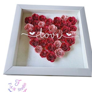 May include: A white framed artwork with a heart made of red and pink paper roses. The word "love" is written across the heart in white script, with heart accents. The background is a light fabric, and the frame is white.