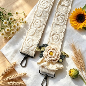 May include: Cream-colored embroidered bag strap with a floral design. The strap features a repeating pattern of roses and leaves. A crocheted flower with green leaves and a cream-colored ruffle is attached to the strap with black metal clasps.