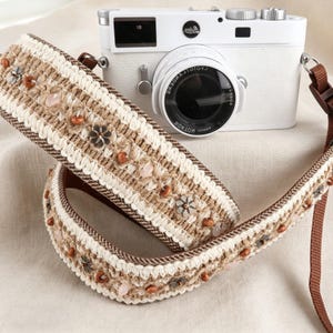 Könnte beinhalten: Eine weiße Kamera mit einem dekorativen Kameragurt. Der Gurt ist beige, braun und weiß, mit floralen und Perlenverzierungen. Der Kameragurt besteht aus gewebtem Material.