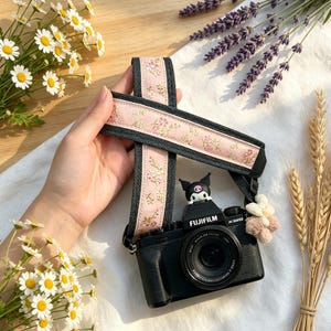 May include: A black Fujifilm X-S20 camera with a pink and grey floral camera strap. The strap features a delicate flower pattern. A small cartoon figure and a flower charm are attached to the camera. Daisies and lavender are in the background.