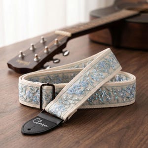May include: A cream guitar strap featuring blue floral embroidery and sequins. The strap has a black leather end with the name "Clara" in cursive. A brown guitar is in the background.