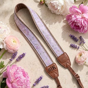 May include: A camera strap with a floral design in lavender and white, with brown leather ends. The strap has the initials "SIK" and a camera icon. The image is decorated with pink and white flowers.