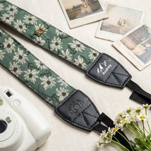 May include: A camera strap featuring a sage green fabric with a white daisy floral pattern. The strap includes black leather-like end tabs, one embossed with a camera icon and the other printed with a mountain graphic and the name Audrey.