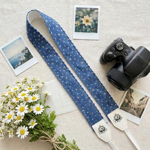 May include: A blue camera strap with a star pattern, featuring white leather ends with a logo. The strap is laid out with a digital camera, polaroid photos, and a bouquet of daisies. The strap is approximately 30 inches long.