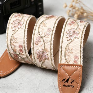 May include: Cream-colored camera strap with embroidered pink floral design and gold accents. The strap has brown leather ends with the name "Audrey" and a mountain graphic. The strap is designed for a camera.
