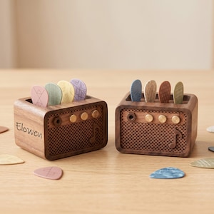 May include: Two wooden guitar pick holders, each resembling a vintage amplifier. They feature speaker grills, brass knobs, and musical notes. The left holder is inscribed with "Elowen". The holders contain colourful guitar picks. The holders are made of wood.