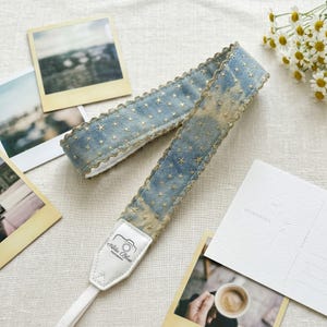 May include: A blue and gold camera strap with a star pattern. The strap has a white leather end piece with the text "Ada Blue Photography" and a camera graphic. The strap is surrounded by polaroid photos and a postcard.