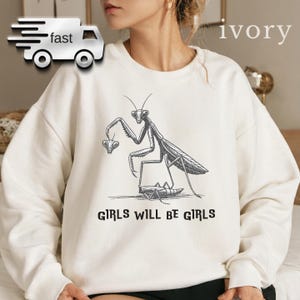 May include: Ivory-colored sweatshirt featuring a black and white illustration of a praying mantis holding a smaller insect. The text "GIRLS WILL BE GIRLS" is printed below the graphic. The sweatshirt has a crew neck and long sleeves.