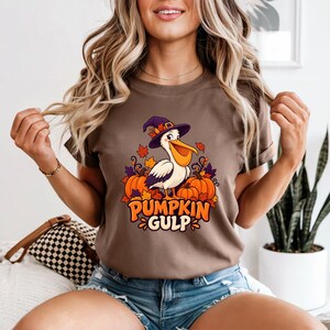 A whimsical illustration of a pelican wearing a purple witch hat, surrounded by orange pumpkins and autumn leaves, with the text PUMPKIN GULP in bold glossy orange letters.