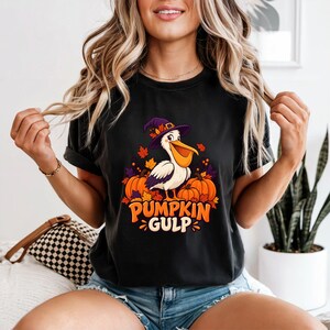 A whimsical illustration of a pelican wearing a purple witch hat, surrounded by orange pumpkins and autumn leaves, with the text PUMPKIN GULP in bold glossy orange letters. — shown on a black product