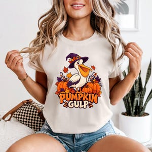 A whimsical illustration of a pelican wearing a purple witch hat, surrounded by orange pumpkins and autumn leaves, with the text PUMPKIN GULP in bold glossy orange letters. — shown on a ivory product
