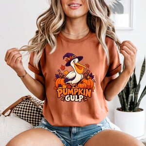 A whimsical illustration of a pelican wearing a purple witch hat, surrounded by orange pumpkins and autumn leaves, with the text PUMPKIN GULP in bold glossy orange letters.