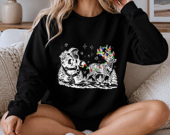 May include: A black long-sleeve sweatshirt featuring a white graphic print of Santa Claus walking with a reindeer. The reindeer is adorned with colourful Christmas lights wrapped around its antlers and body, set against a snowy forest background.