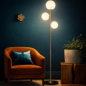 May include: A modern floor lamp with three spherical, frosted glass shades. The lamp has a gold-colored metal frame and a black base. An orange armchair with a teal pillow sits nearby. A potted plant and wooden side table complete the scene.