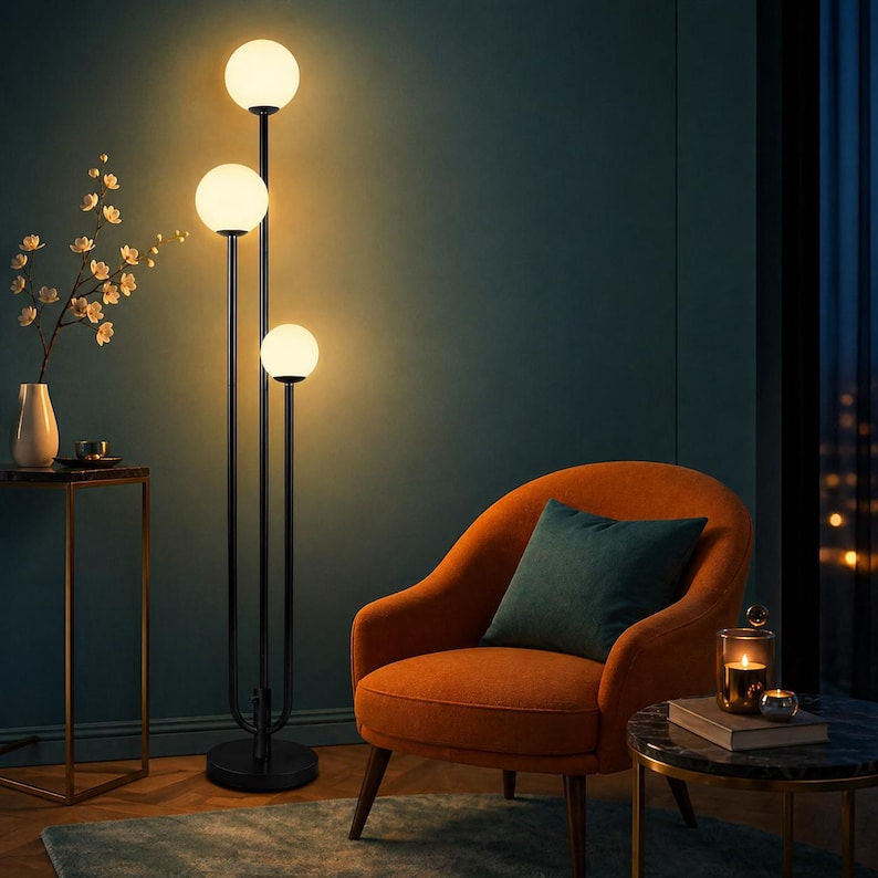 May include: A modern black floor lamp with three spherical, glowing white lights. The lamp stands tall next to an orange armchair with a teal cushion. The room has a dark teal wall and a small side table with a vase of flowers.